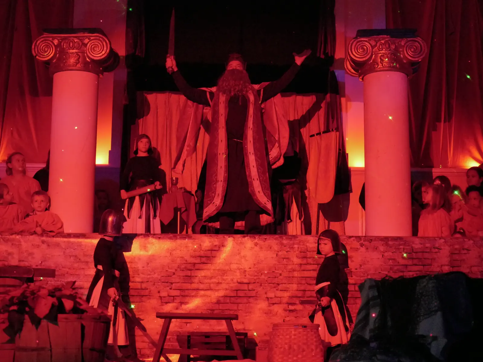 The king raising a sword between two columns in red light, with child soldiers below