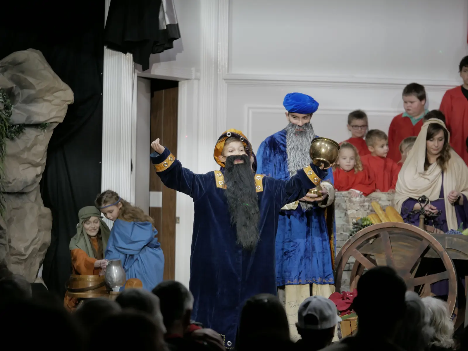 A young wise man in blue velvet and a black beard, arms wide, holding a gold cup